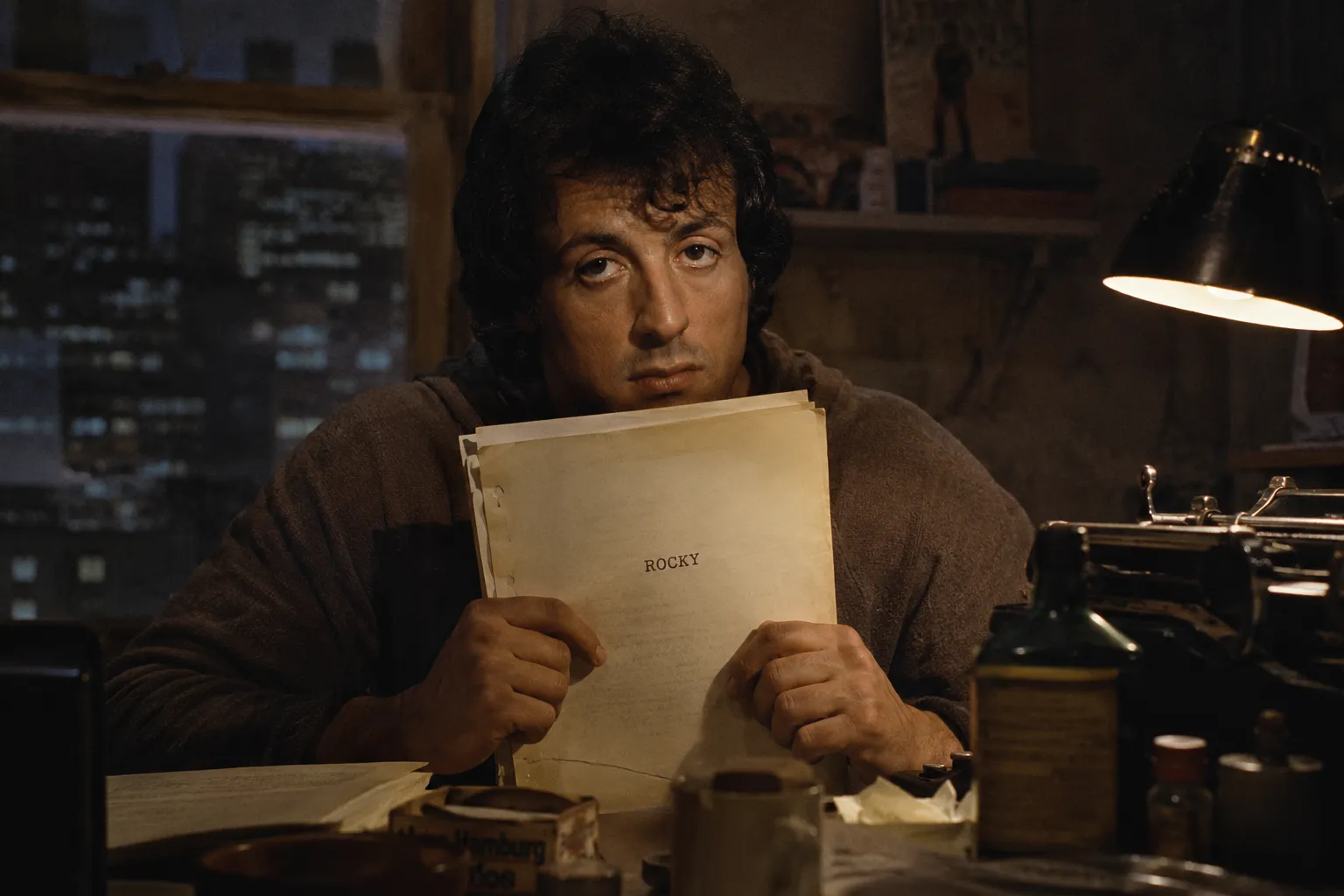 Sylvester Stallone sosteniendo el guion de Rocky frente a una máquina de escribir